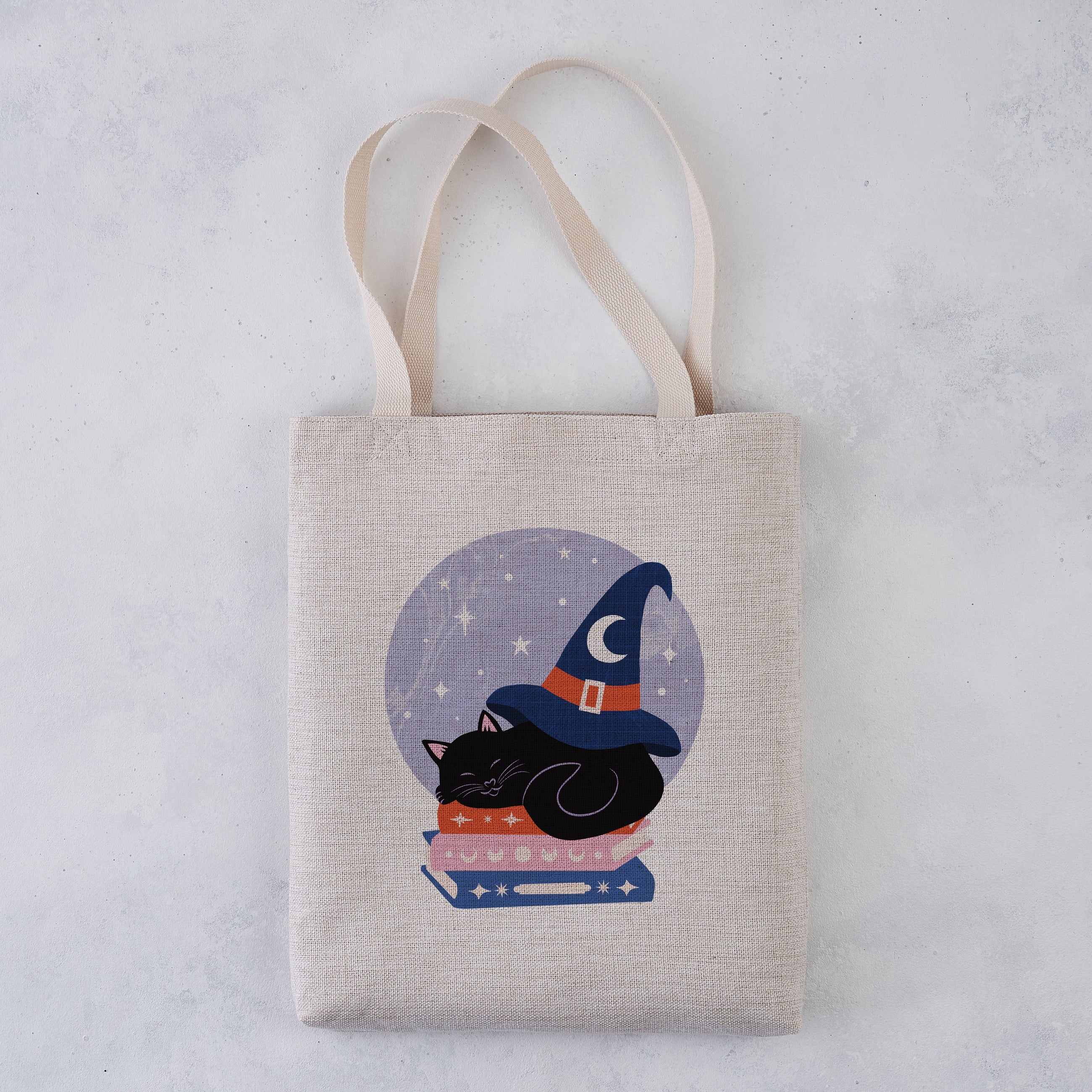 A natural canvas tote bag with a printed illustration of a black cat wearing a wizard hat on top of books inside a circular moonlit design, shown against a light gray textured background.