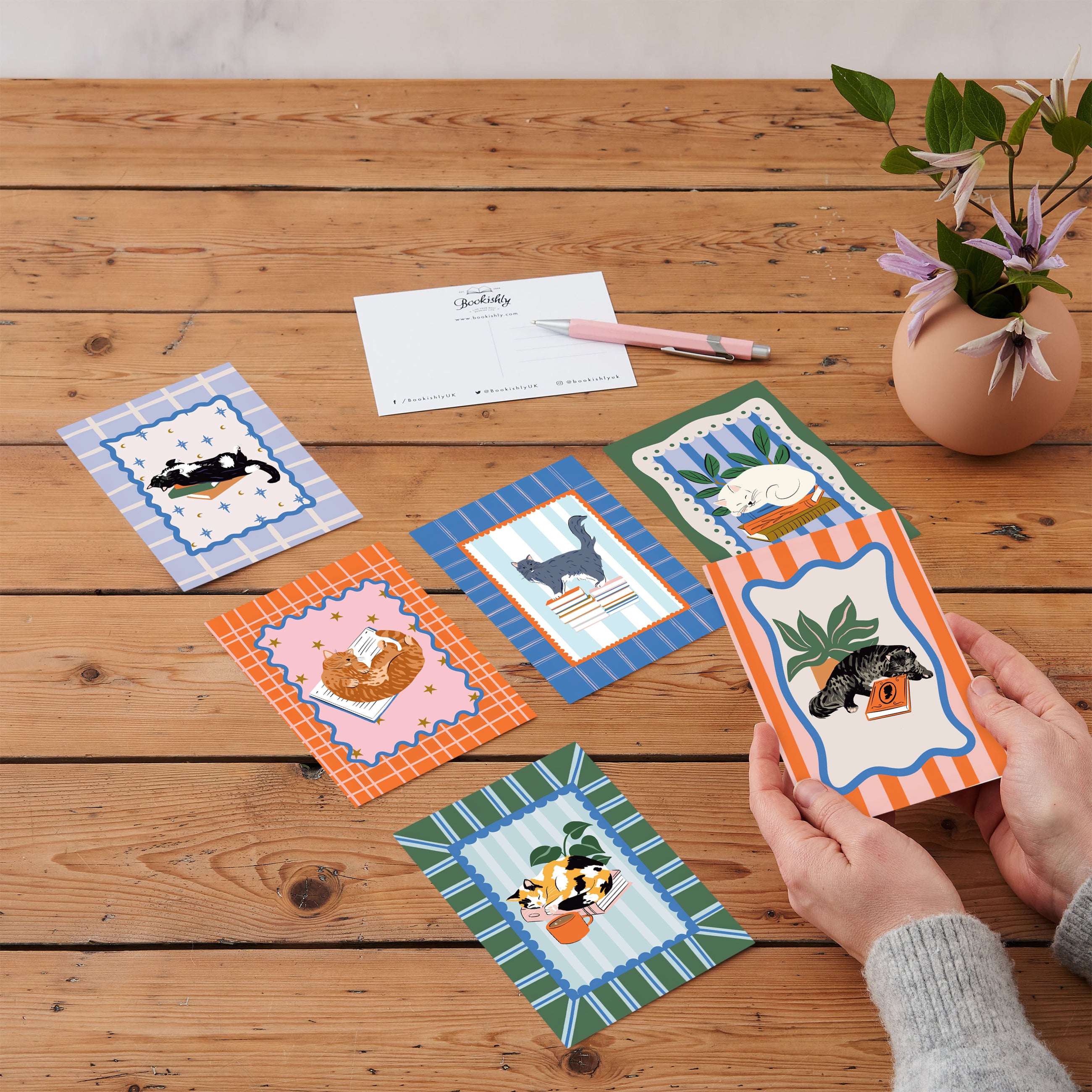 A set of six postcards with various illustrations including cats and books displayed on a wooden table. One postcard is being held by a hand, showing a colorful design with a pink background and decorative elements.