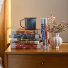 Stack of books and a mug on a wooden nightstand with a vase of flowers.