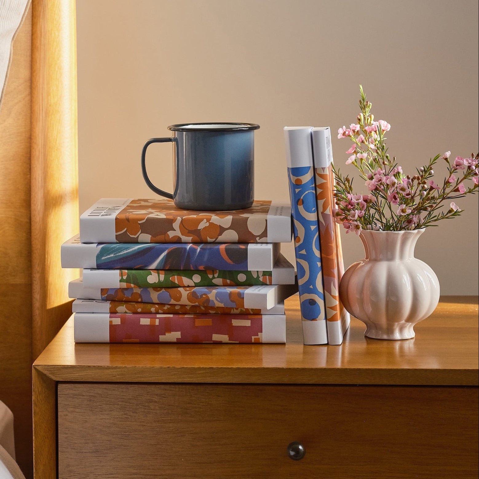 Stack of books and a mug on a wooden nightstand with a vase of flowers.