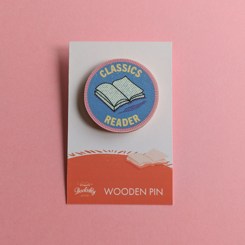 Classics Reader wooden pin on a pink background, featuring an open book on a round blue badge with pink edging.