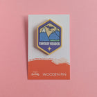 Fantasy Reader wooden pin on a pink background, featuring a purple and gold badge with a starry mountain design.