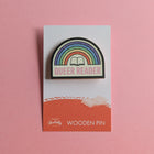 Queer Reader wooden pin on a pink background, featuring a stitched rainbow design with an open book and pink lettering.