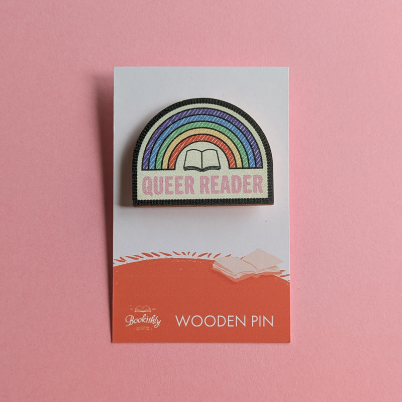 Queer Reader wooden pin on a pink background, featuring a stitched rainbow design with an open book and pink lettering.