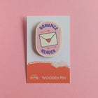 Romance Reader wooden pin on a pink background, featuring a love letter and arrows on a pink badge design.