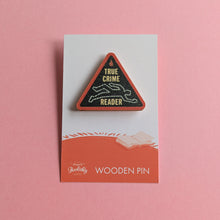 True Crime Reader wooden pin on a pink background, shaped like a red warning triangle with a chalk outline graphic.