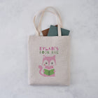 Personalised book bag tote with a pink cat holding a green book and the text “Dylan’s Book Bag” printed above.