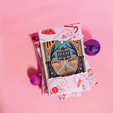 Wrapped book advent calendar with Emily Brontë edition partly unwrapped, surrounded by pink and purple baubles on a pink background.