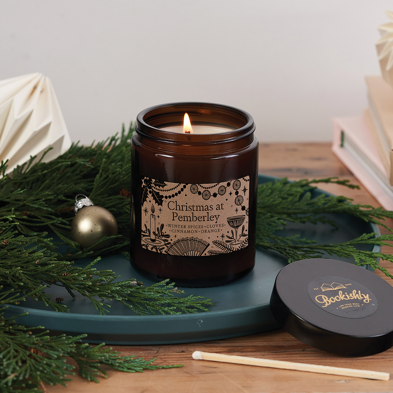 Brown glass candle jar labelled “Christmas at Pemberley” surrounded by festive greenery, books, and a matchstick – a cosy literary candle gift for readers
