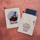 Two cream-colored canvas sleeve pouches with black zipper pulls, displayed on a textured pink background. The sleeve on the left features a graphic of a black cat in a witch hat on books inside a starry circle.