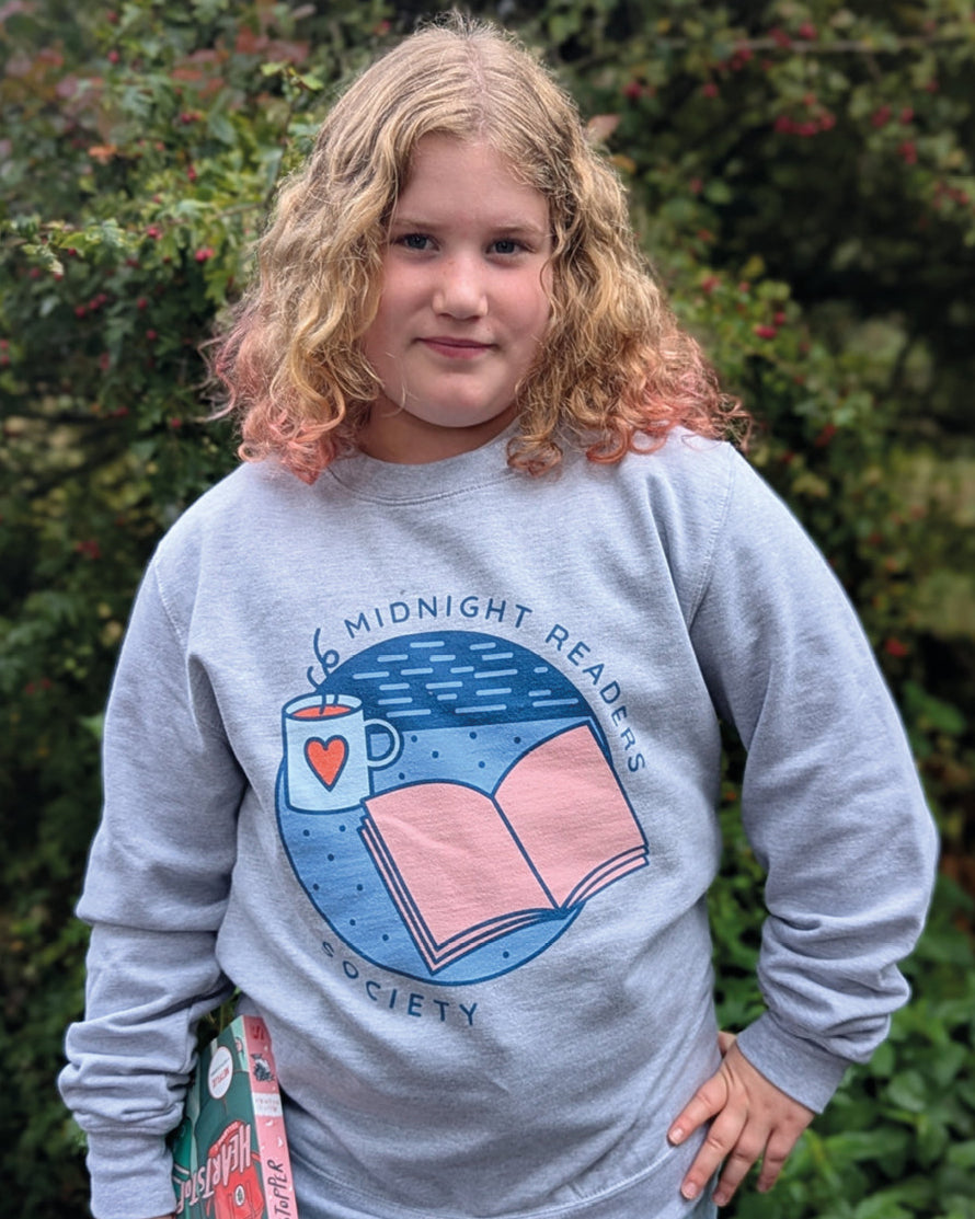 Person wearing a sweatshirt with a book and cup design, standing in front of green foliage.