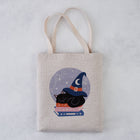 A natural canvas tote bag with a printed illustration of a black cat wearing a wizard hat on top of books inside a circular moonlit design, shown against a light gray textured background.