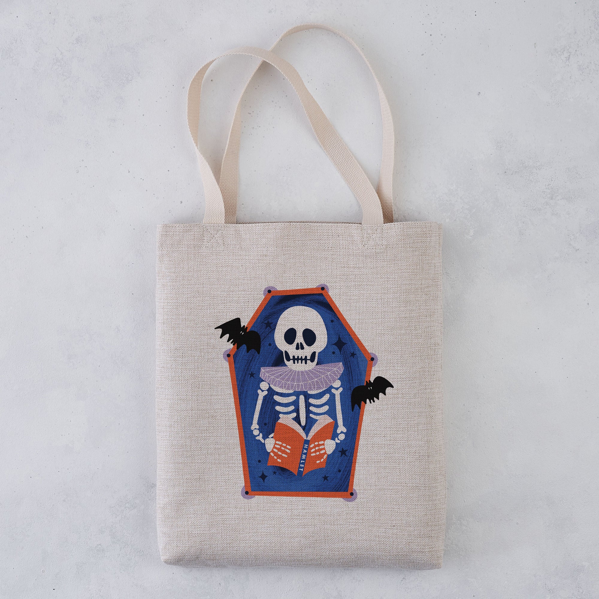 A light beige canvas tote bag with two shoulder straps, featuring a printed coffin-shaped graphic of a skeleton reading an orange book with bats on a blue background, set against a light gray textured backdrop.