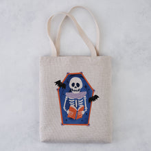 A light beige canvas tote bag with two shoulder straps, featuring a printed coffin-shaped graphic of a skeleton reading an orange book with bats on a blue background, set against a light gray textured backdrop.