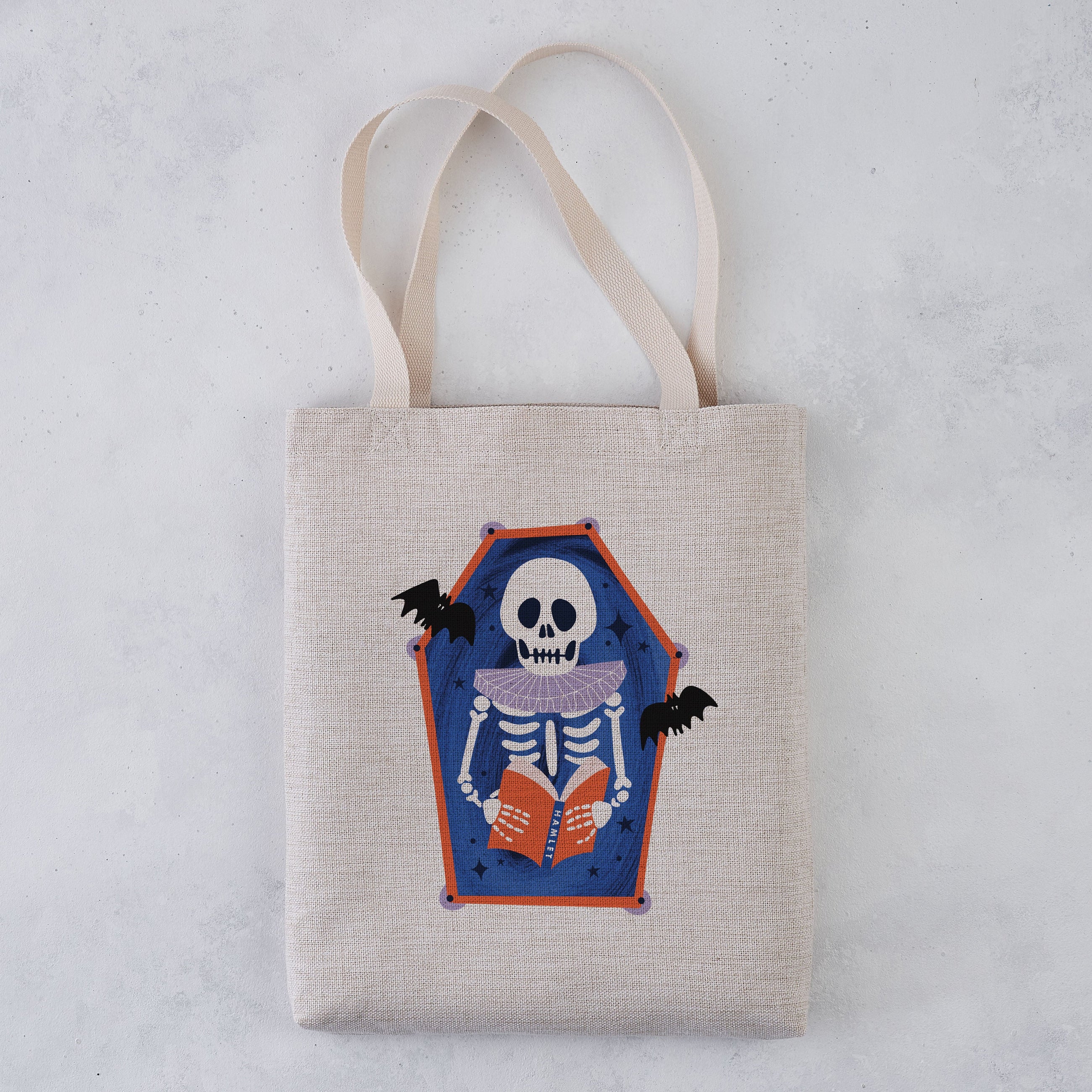 A light beige canvas tote bag with two shoulder straps, featuring a printed coffin-shaped graphic of a skeleton reading an orange book with bats on a blue background, set against a light gray textured backdrop.