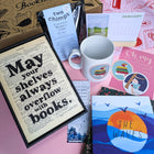 A winter-themed gift box for readers displayed on a pink background, with a framed book page print, classic novel, festive mug, speciality coffee, 2026 postcard calendar, bookmarks, postcards and seasonal stickers.