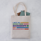 A beige tote bag with the text 'I BUY MY BOOKS FROM BOOKSHOPS NOT BILLIONAIRES' printed in multicolored letters. The bag is filled with books and is placed on a light-colored surface.