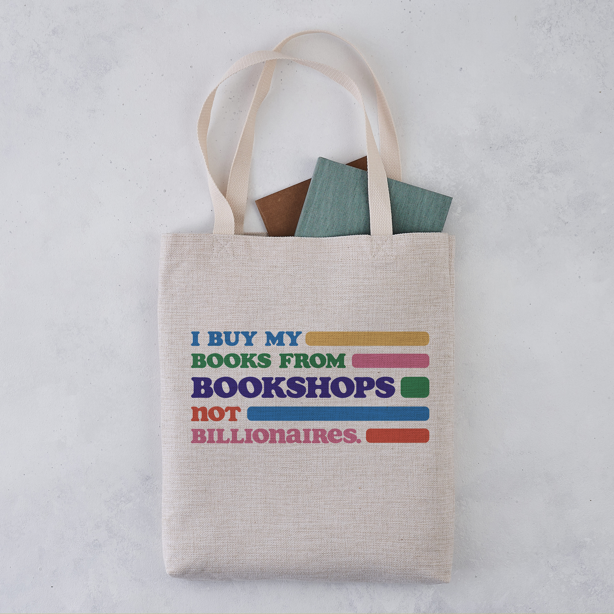 A beige tote bag with the text 'I BUY MY BOOKS FROM BOOKSHOPS NOT BILLIONAIRES' printed in multicolored letters. The bag is filled with books and is placed on a light-colored surface.
