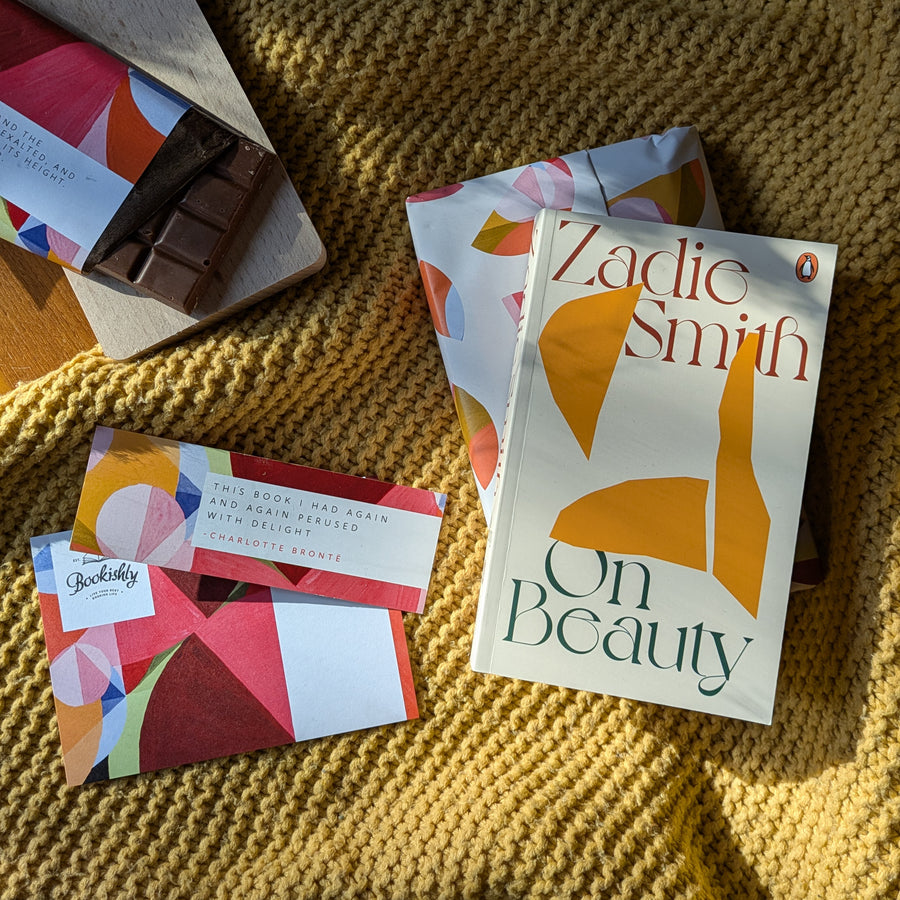 Flat lay of Bookishly's Classics Revisited subscription showing a monthly bundle featuring Zadie Smith's 'On Beauty' with a colourful matching chocolate bar and coordinated wrapping. The book, chocolate, and packaging rest on a mustard yellow knitted blanket with sunlight across the scene.