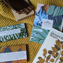 Flat lay of Bookishly's Modern Classics subscription showing two monthly bundles. Includes 'The Sea, The Sea' by Iris Murdoch and 'Giovanni's Room' by James Baldwin, each paired with matching illustrated chocolate bars. Everything is arranged on a mustard yellow knitted blanket with one chocolate bar partly unwrapped.