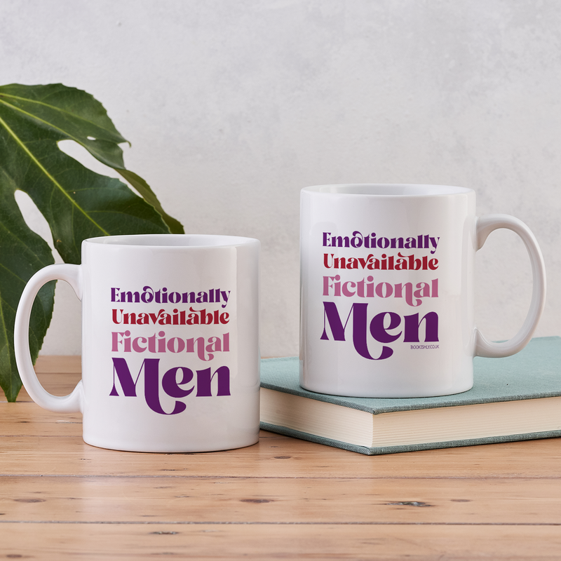 White ceramic mug with the phrase “Emotionally Unavailable Fictional Men” in red and purple lettering, styled on a wooden table beside a green leaf and a book.