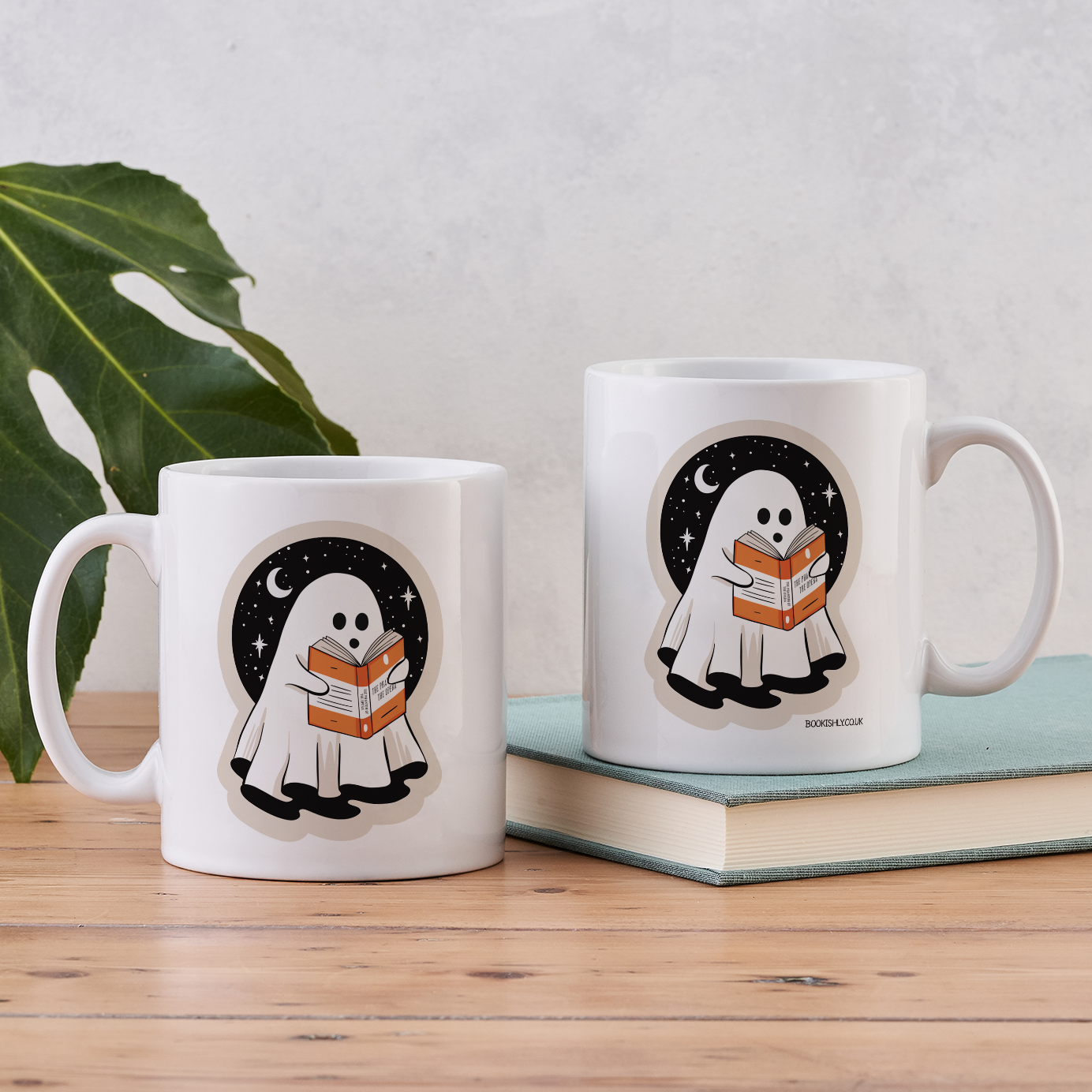 Two white ghost mugs with reading design, displayed on wooden table with plant and book.
