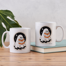 Two white ghost mugs with reading design, displayed on wooden table with plant and book.