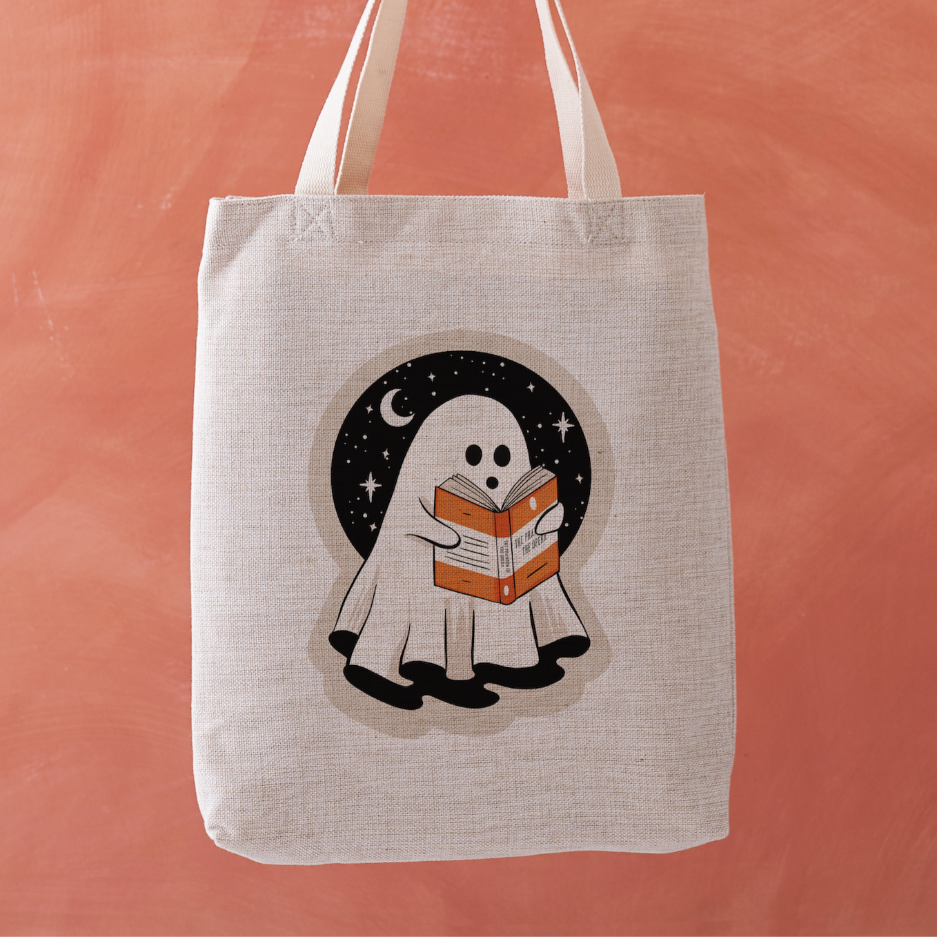 Linen-effect tote bag with ghost reading a book design, held against coral background.