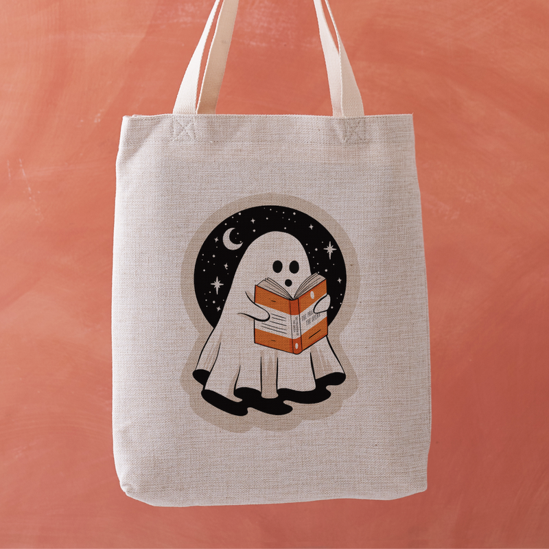 Linen-effect tote bag with ghost reading a book design, held against coral background.