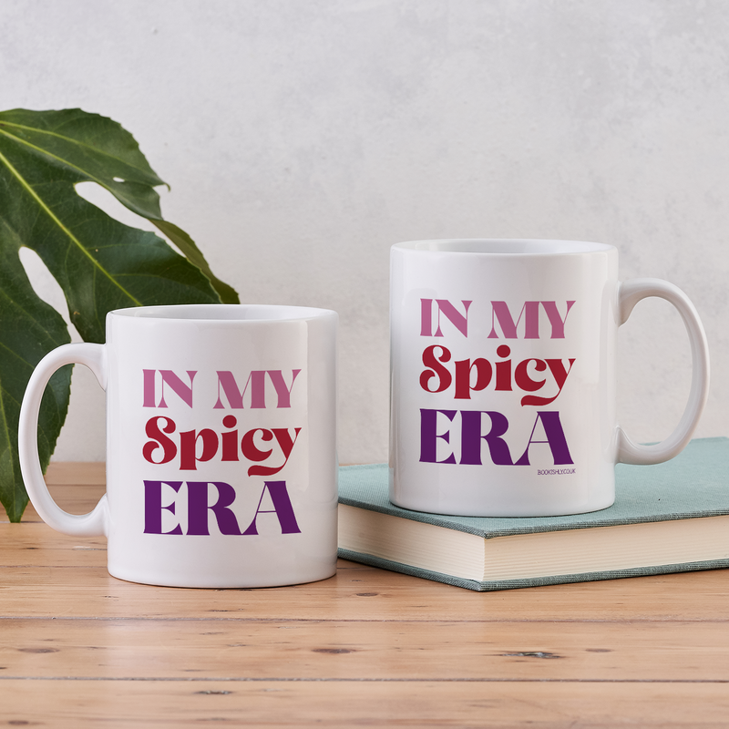 White ceramic mug with the phrase “In My Spicy Era” in red and purple lettering, styled on a wooden table beside a green leaf.