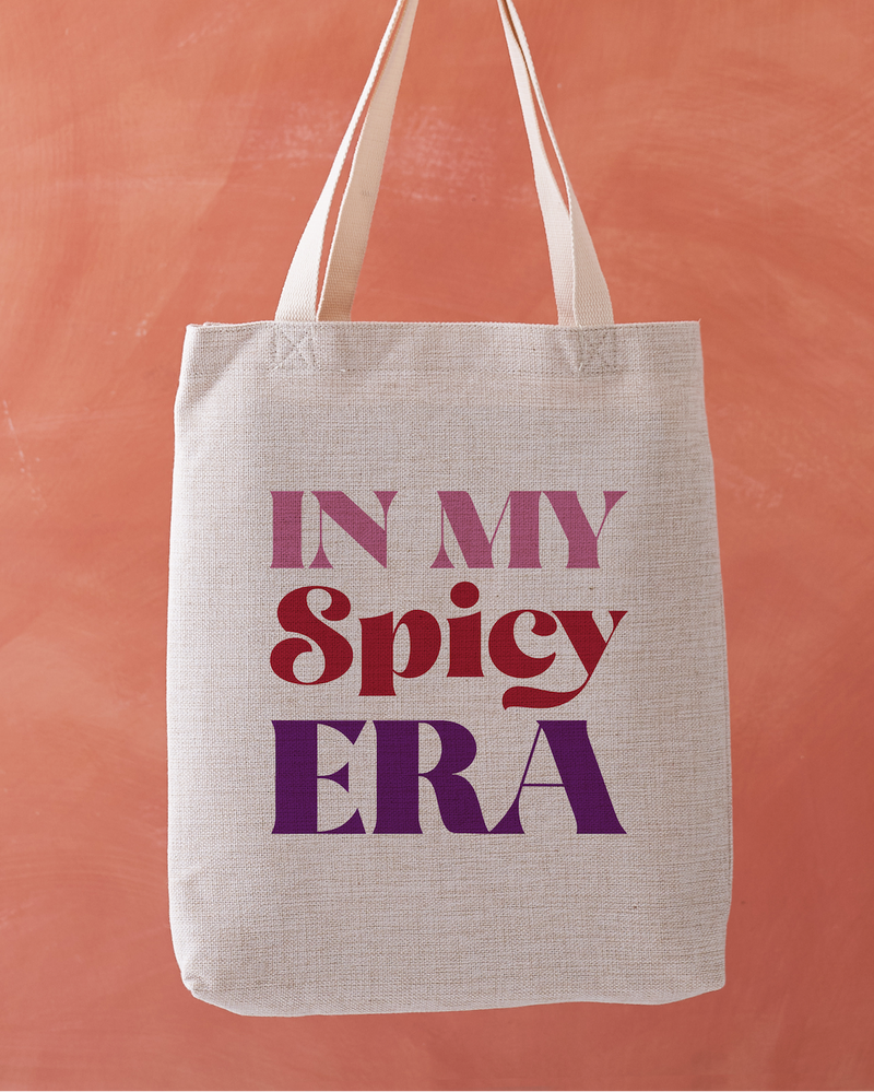 Linen-effect tote bag with the phrase “In My Spicy Era” in red and purple lettering, held against a warm terracotta background.