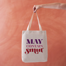 Linen-effect tote bag with the phrase “May Contain Smut” in red and purple lettering, held against a warm terracotta background.