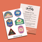 A sheet of six colourful temporary tattoos featuring genre reader designs – Queer Reader (rainbow), Classics Reader (open book), Fantasy Reader (mountains), YA Reader (comic cloud), Romance Reader (love letter), and True Crime Reader (handprint). Includes an instruction card on a terracotta background.