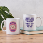 White ceramic mug with “Romance Reader” patch design on one side and a list of romance tropes on the other, styled on a wooden table beside a green leaf.