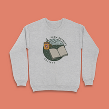Grey sweatshirt with “Slow Readers Society” design featuring an open book, steaming mug with heart, and illustrated night sky.