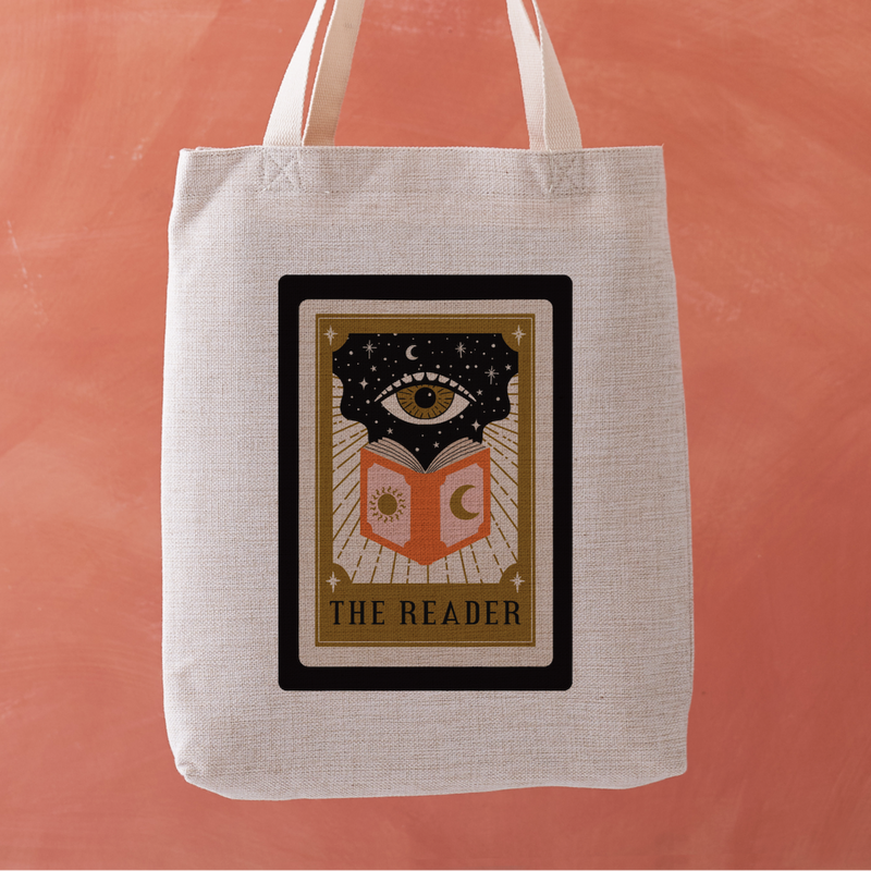 Linen-effect tote bag with tarot card design reading “The Reader,” held against coral background.