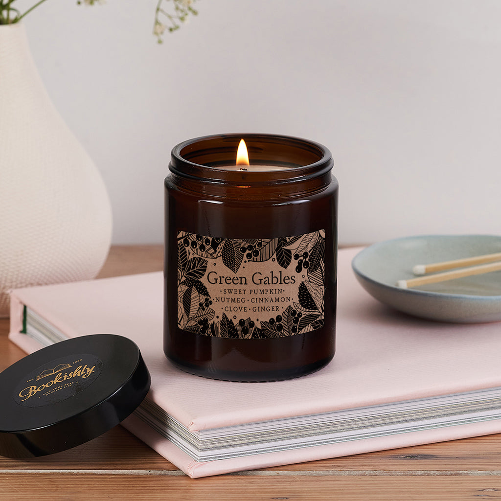 Green Gables candle in brown glass jar with leafy kraft label, lit and sitting on a pink book beside a black Bookishly lid