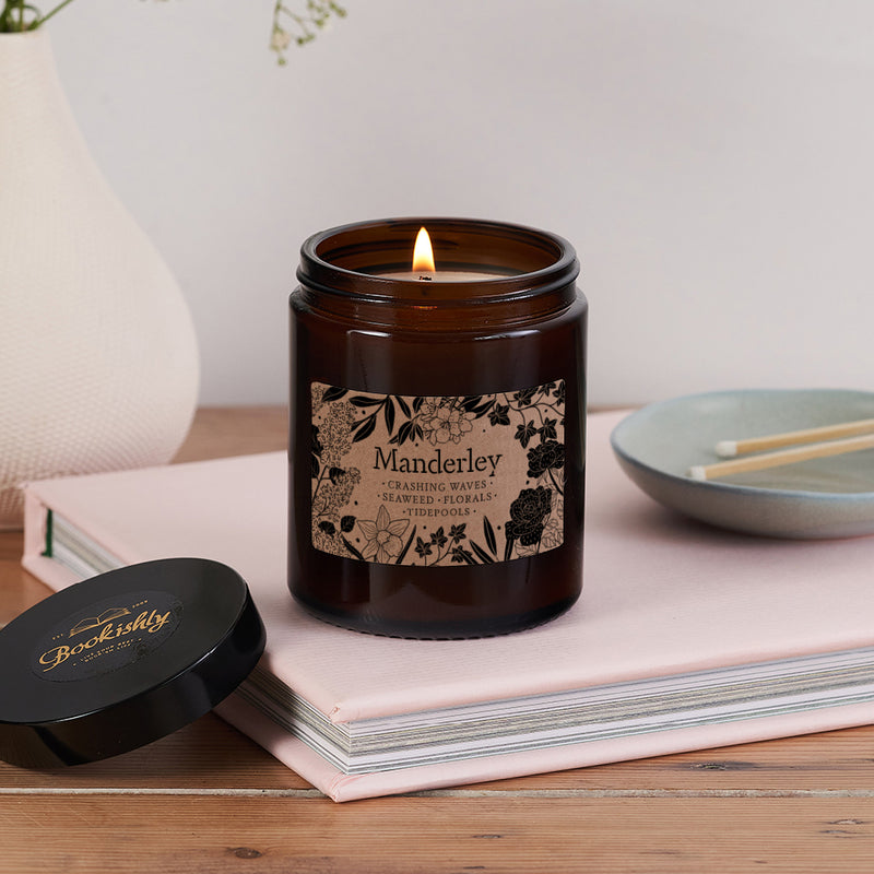 Lit Manderley candle in brown glass jar with floral kraft label, placed on pink book with lid beside it