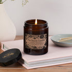 The Secret Garden candle in a dark amber glass jar with floral kraft label, lit and resting on a pale pink book beside a Bookishly lid and ceramic match dish