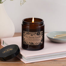 The Secret Garden candle in a dark amber glass jar with floral kraft label, lit and resting on a pale pink book beside a Bookishly lid and ceramic match dish