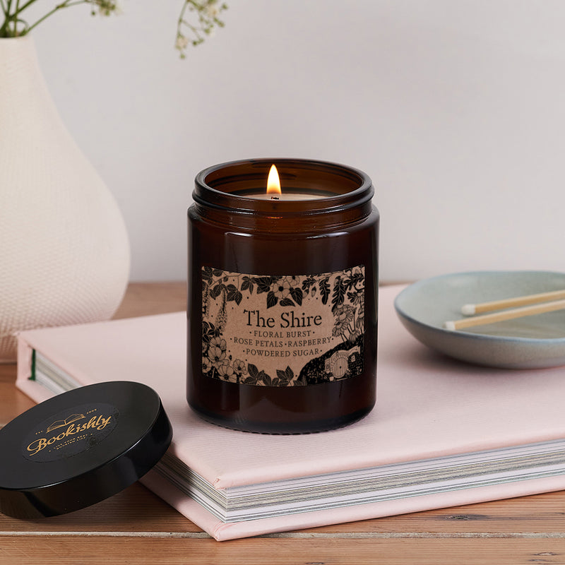 The Shire candle in brown glass jar with floral kraft label, lit and placed on a pink book with a black Bookishly lid beside it