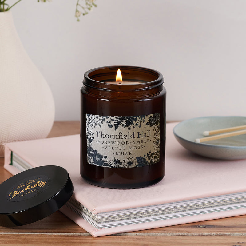 Thornfield Hall candle in amber glass jar with gothic floral label, lit and resting on a pale pink book next to Bookishly lid and match dish